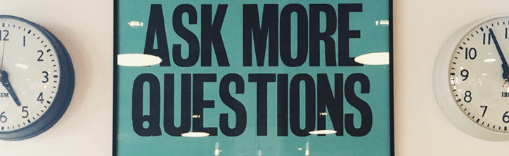 Framed sign reading ‘Ask More Questions’ mounted on a wall between two clocks.