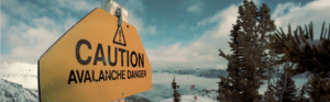 Yellow caution sign reading &lsquo;Caution: Avalanche Danger&rsquo; positioned on a snowy mountain landscape.