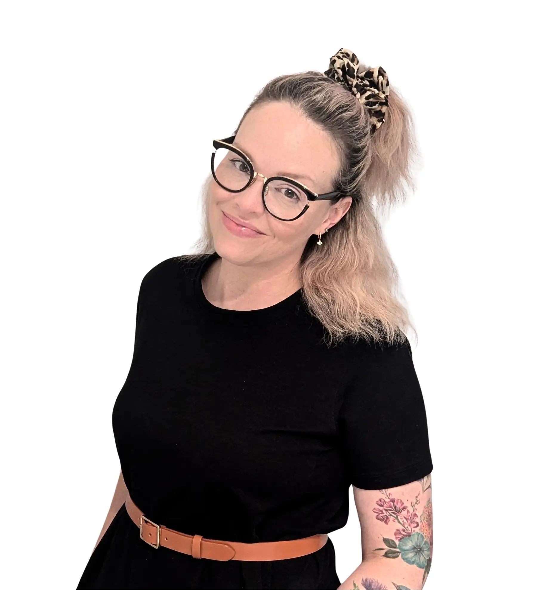 A woman with blonde hair pulled back with a leopard-print scrunchie, wearing glasses and a black dress with a brown belt, smiling slightly and standing against a white background.