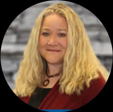 A woman with long wavy blonde hair wearing a maroon blazer and black top, smiling softly in front of a blurred gray background.
