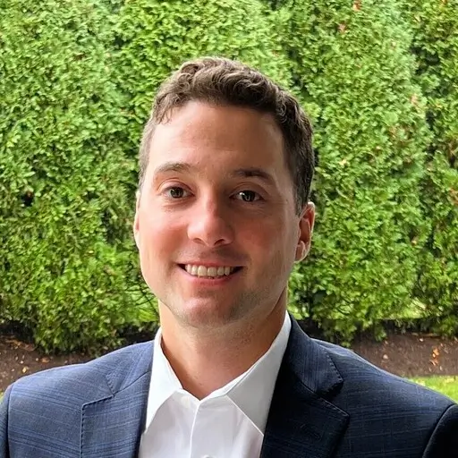 A man with short brown hair wearing a dark blue suit, smiling while standing outdoors.